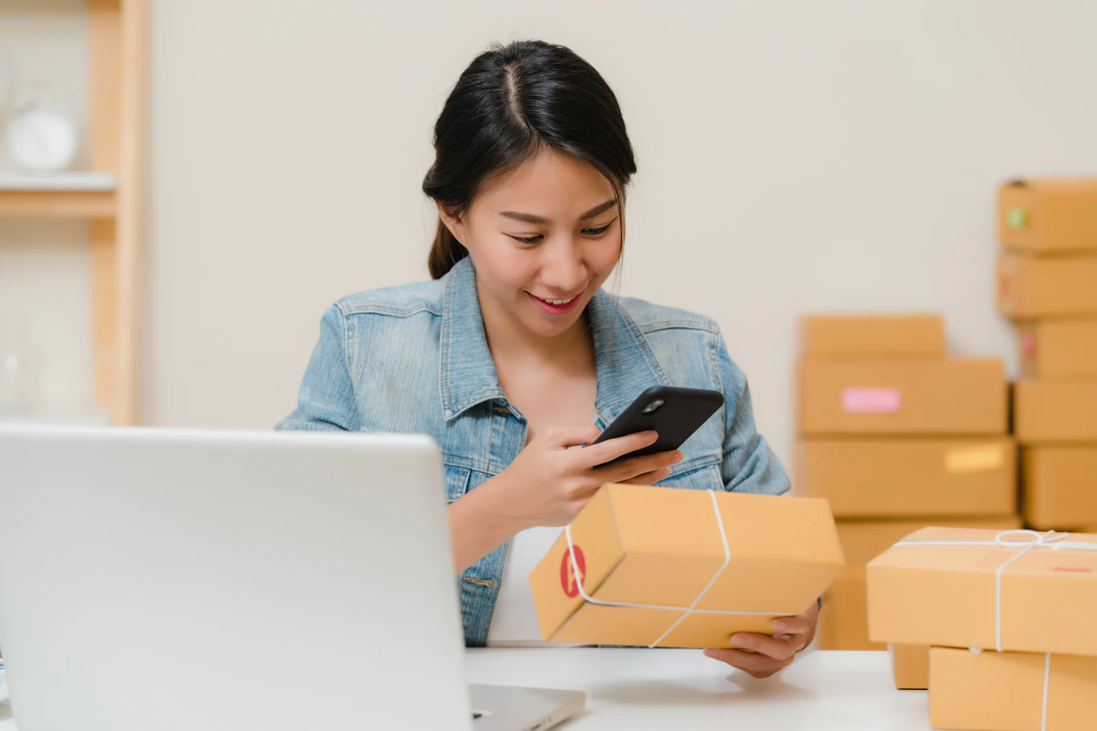 Frau prüft ein Paket mit dem Smartphone, daneben Laptop und Versandkartons